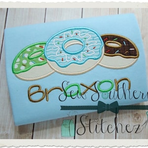 May include: A light blue t-shirt with three embroidered donuts. The donuts are green with white sprinkles, blue with white sprinkles and brown with chocolate sprinkles. The text "Braxton Southern Stitchez" is embroidered below the donuts.