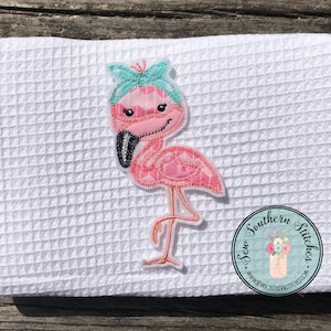 May include: White waffle-weave towel with a pink flamingo applique. The flamingo has a teal headband and is standing on one leg. The applique is stitched with pink thread and has a black beak. A round logo is in the bottom right corner.