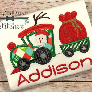 May include: Red and green Christmas train with Santa Claus driving and a red sack of gifts in the back. The train is on a white shirt with the name Addison embroidered in red.