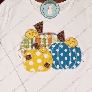 Puede incluir: Una camiseta blanca con un diseño de tres calabazas en patchwork. Las calabazas son amarillas, azules y verdes con lunares blancos. Las calabazas están rodeadas de hojas marrones.