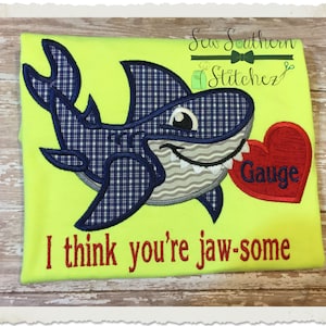 May include: A blue and white plaid shark holding a red heart with the name "Gauge" embroidered on it. The text "I think you're jaw-some" is embroidered below the shark.
