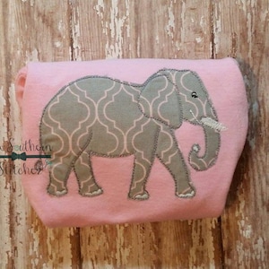 Könnte beinhalten: Ein rosa T-Shirt mit einem gestickten grauen Elefantenmotiv. Der Elefant hat ein geometrisches Muster und ein kleines Augendetail.