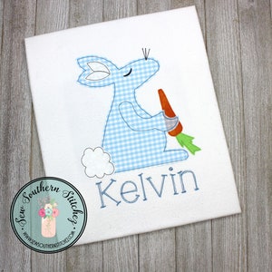 May include: A white onesie with a blue and white gingham bunny holding a carrot. The bunny is sitting down and the onesie is personalized with the name "Kelvin".
