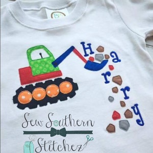Puede incluir: Una camiseta blanca con un diseño bordado de una excavadora de juguete verde y roja excavando rocas marrones y grises. El texto "Happy" está bordado en azul sobre la excavadora. El texto "Sew Southern Stitchez" está bordado en negro debajo de la excavadora.