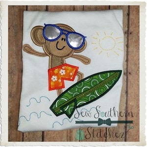 Puede incluir: Diseño bordado de un mono con gafas de sol y pantalones cortos, montando una tabla de surf con rayas verdes y blancas, con un sol y olas de fondo. El diseño está en una camiseta blanca.