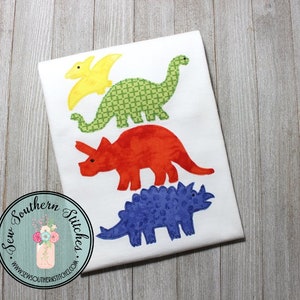 May include: A white fabric with three embroidered dinosaurs. The dinosaurs are a yellow and green pterodactyl, a red triceratops, and a blue stegosaurus.
