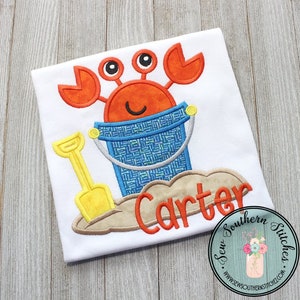 May include: A white t-shirt with an embroidered design of a red crab holding a blue bucket and a yellow shovel. The name "Carter" is embroidered in orange below the crab.