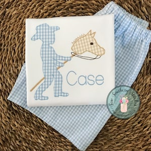 May include: A white onesie with a blue gingham pattern, featuring a boy holding a stick with a gingham horse head attached. The onesie is personalized with the name "Case".