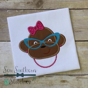 Puede incluir: Diseño bordado de un mono marrón con gafas azules y un lazo rosa. El mono lleva un collar de cuentas rosas. El diseño está en una camiseta blanca.