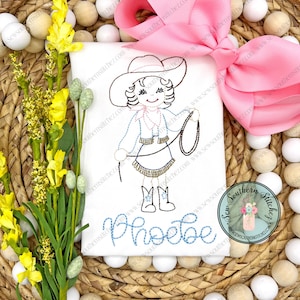 May include: A white t-shirt with a brown embroidered image of a girl wearing a cowboy hat and holding a lasso. The girl's name, "Phoebe", is embroidered in blue below the image. The t-shirt is surrounded by yellow flowers and a pink bow.