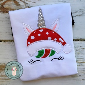 May include: White t-shirt with an embroidered unicorn wearing a red and white Santa hat with white polka dots. The unicorn has a silver horn, pink ears, and green and red mane.