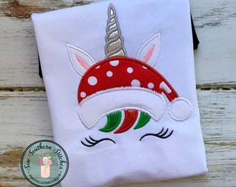 May include: White t-shirt with an embroidered unicorn wearing a red and white Santa hat with white polka dots. The unicorn has a silver horn, pink ears, and green and red mane.
