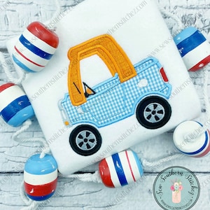 Peut inclure: Un camion en tissu gingham bleu et blanc avec une cabine orange. Le camion est brodé sur un tissu blanc avec une bordure en corde et des bouées nautiques en corde rouge, blanche et bleue.