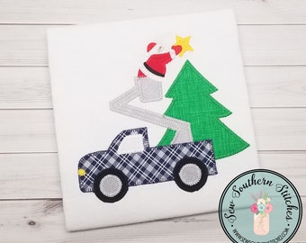 May include: A white t-shirt with an applique of a truck with a lift, Santa Claus, and a Christmas tree. The truck is blue and white plaid, the lift is gray, Santa is red and white, and the tree is green. The text "Sew Southern Stitches" is on the bottom right corner of the shirt.