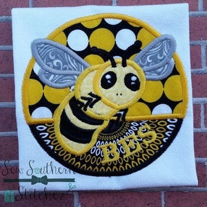 May include: A yellow and black bee with a gray and white patterned wing is embroidered on a yellow and black polka dot circle. The circle has a black border and the word "Bees" is embroidered in yellow letters on the bottom.