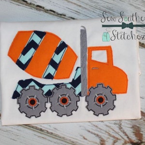 Puede incluir: Una camiseta blanca con una aplicación naranja y azul de un camión mezclador de cemento. El camión tiene un patrón de chevron en el tambor y engranajes grises para las ruedas. El texto "Sew Southern Stitchez" está en la esquina superior derecha de la imagen.