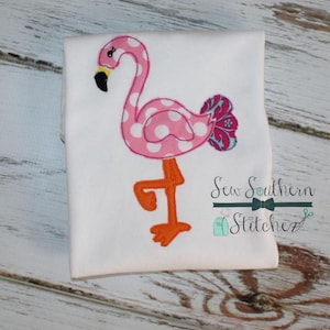 May include: Pink flamingo with white polka dots and a blue and pink ruffled tail. The flamingo is standing on one leg with an orange leg and foot. The image is on a white t-shirt with the text "Sew Southern Stitchez" in black.