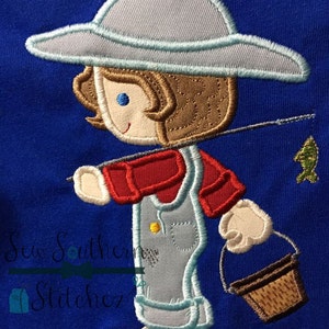 Può includere: Design ricamato di un pescatore di cartoni animati con cappello blu e tuta, che tiene una canna da pesca con un pesce e un piccolo cestino. Il pescatore indossa una camicia rossa e una tuta azzurra. Lo sfondo è blu.