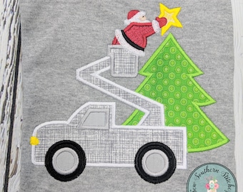 May include: Santa Claus in a red suit and black belt is in a bucket truck, placing a yellow star on top of a green Christmas tree with white polka dots. The truck is white with black tires and a gray cab.