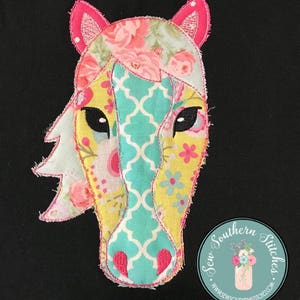 Op de afbeelding: Een kleurrijke patchwork-paardenkop-applicatie met roze oren en een turquoise en gele gepatroneerde neus. De applicatie is genaaid op een zwarte stoffen achtergrond. De tekst "Sew Southern Stitches" is zichtbaar in een cirkel onder de paardenkop.