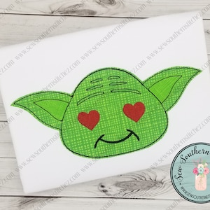 May include: A white t-shirt featuring a green cartoon character's face with heart-shaped eyes and a smile. The character has large ears and is embroidered with a crosshatch pattern. The design is centered on the shirt.