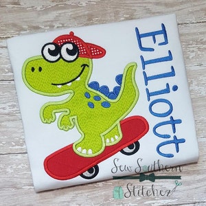 Puede incluir: Una camiseta blanca con un dinosaurio verde bordado que lleva una gorra roja y blanca y monta un monopatín rojo. El dinosaurio tiene ojos azules y una cola azul. El texto "Elliott" está bordado en azul en el lado derecho de la camiseta.