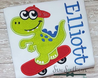 May include: A white t-shirt with an embroidered green dinosaur wearing a red and white baseball cap and riding a red skateboard. The dinosaur has blue eyes and a blue tail. The text "Elliott" is embroidered in blue on the right side of the shirt.