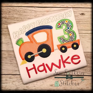 May include: A white t-shirt with an embroidered design of a red and orange train pulling a wagon with the number 3 on it. The text "Happy Birthday" is above the train and the name "Hawke" is below the train.