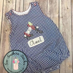 May include: A blue and white gingham baby romper with a decorative apple and ant design. The romper has a white collar and red buttons. The name "Axel" is embroidered on the front.