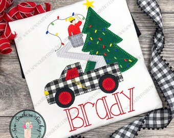 May include: White t-shirt with an embroidered design featuring a black and white plaid truck carrying a Christmas tree. A person is putting lights on the tree. The name "Brady" is embroidered in red.