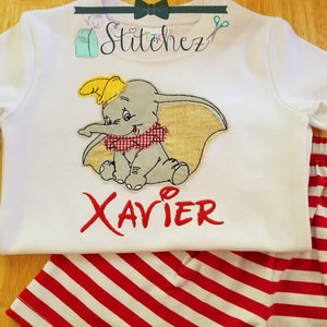 Puede incluir: Camiseta blanca de manga corta con una imagen bordada de Dumbo el elefante, que lleva una bufanda a cuadros rojos y blancos. El nombre "XAVIER" está bordado en rojo debajo de la imagen. La camiseta se combina con pantalones cortos de rayas rojas y blancas.