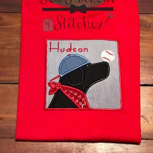 May include: Red t-shirt with an embroidered design of a black dog wearing a blue baseball cap and a red bandana. The dog is looking to the right and there is a baseball to the right of the dog. The text "Hudson" is above the dog.