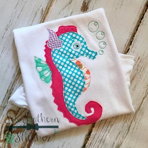 May include: A white t-shirt with a pink, teal, and green appliqué of a seahorse with white polka dots. The seahorse has a pink fin and a green fin with white polka dots. There are three green circles above the seahorse.