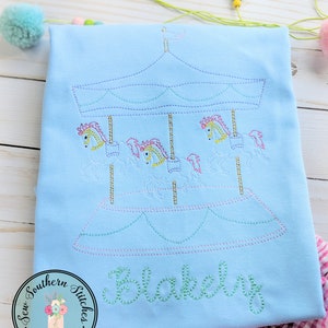 May include: A light blue t-shirt with an embroidered carousel design and the name "Blakely" stitched in green thread.