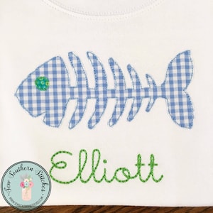 May include: A white t-shirt with a blue and white gingham fish bone applique and the name "Elliott" embroidered in green thread.