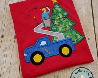 May include: A blue toy truck with a lift is decorated with a green Christmas tree with lights and a star on top. The truck is on a red background.