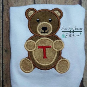 May include: Brown teddy bear applique with a brown circle and the letter T in red. The applique is on a white shirt. The text "Sew Southern Stitchez" is in the top right corner of the image.