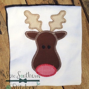 May include: A white t-shirt with a brown reindeer applique. The reindeer has a red and white checked nose.
