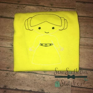 May include: A yellow t-shirt with an embroidered image of Princess Leia from Star Wars. The embroidery is in black thread and shows a cartoon-style image of Princess Leia with her signature hair buns and a belt. The text "Sew Southern Stitchez" is embroidered in black thread at the bottom of the shirt.