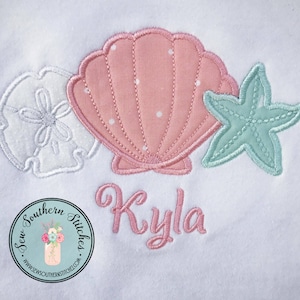 May include: A white onesie with a pink seashell, a green starfish, and a white seashell. The onesie is embroidered with the name "Kyla".
