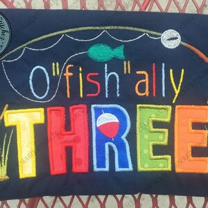 May include: A blue embroidered shirt with the text "O'fish'ally Three" in colorful letters. The design includes a fishing rod, a fish, and a bobber.