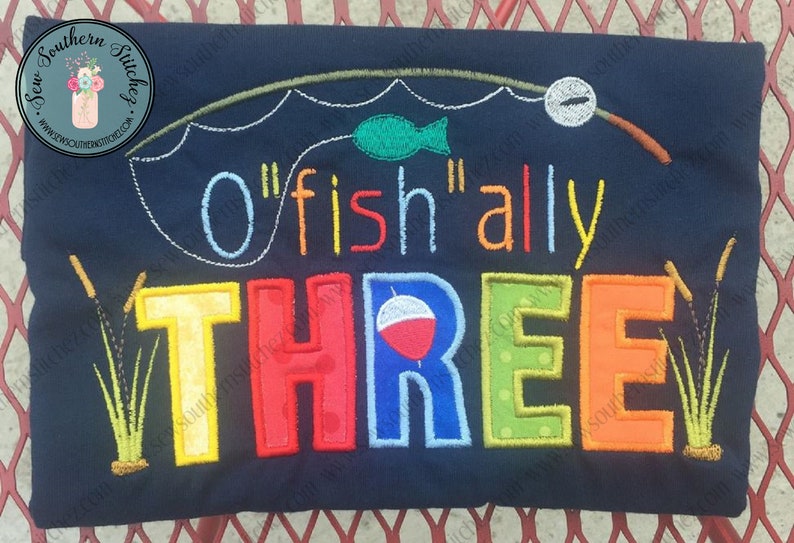 May include: A blue embroidered shirt with the text "O'fish'ally Three" in colorful letters. The design includes a fishing rod, a fish, and a bobber.