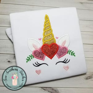 Puede incluir: Camiseta blanca con un diseño de unicornio bordado. El unicornio tiene un cuerno dorado, orejas rosas y tres corazones rojos, rosas y brillantes. El unicornio también tiene pestañas y un pequeño corazón rosa debajo de cada ojo.