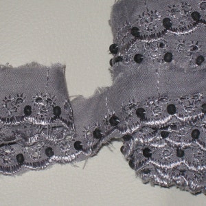 May include: Grey lace trim with black beads. The trim has a scalloped edge and a delicate floral pattern.