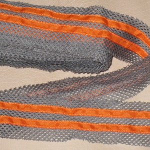 May include: A roll of grey mesh ribbon featuring two parallel orange satin stripes. The ribbon has an open, textured weave, suitable for crafts or decorative use. The orange stripes provide a vibrant contrast against the grey background.