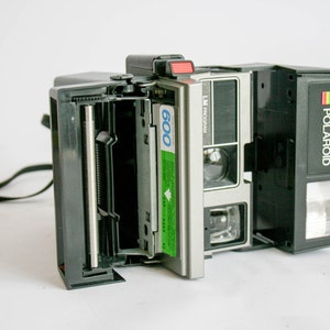 May include: A black Polaroid Land Camera with a silver front and a green 600 film pack inserted. The camera has a built-in flash and a black strap.