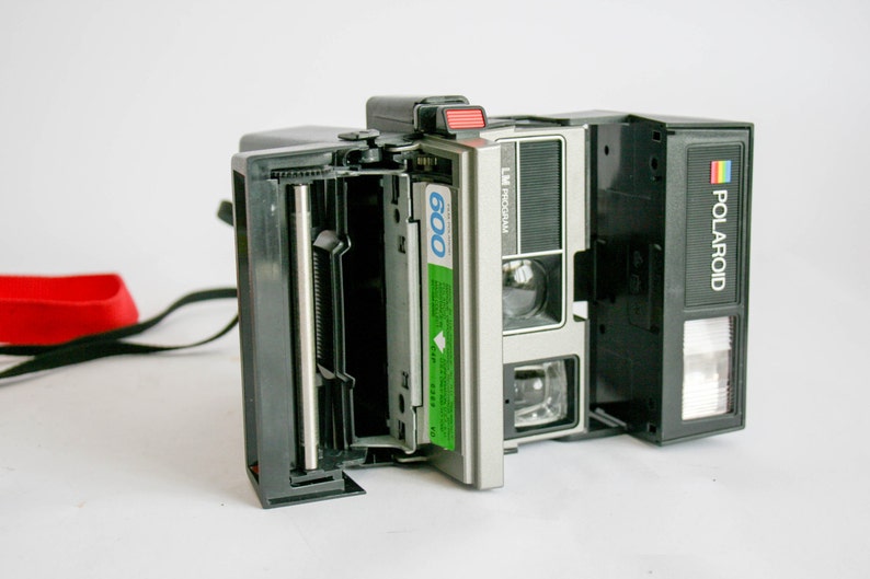 May include: A black Polaroid Land Camera with a silver front and a green 600 film pack inserted. The camera has a built-in flash and a black strap.
