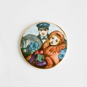 May include: A round porcelain ornament with a gold trim features a painted image of a couple in winter attire. The man is wearing a blue uniform and a hat, while the woman is wearing a red coat and holding a wrapped gift. A white horse is partially visible in the background.