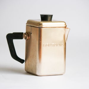 K&ouml;nnte beinhalten: Eine goldene Kaffeekanne aus Metall mit einem schwarzen Kunststoffgriff und -deckel. Die Kanne tr&auml;gt die Aufschrift "MONTANA" auf der Seite.