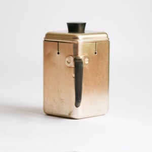 K&ouml;nnte beinhalten: Ein goldfarbener Metallbeh&auml;lter mit einem schwarzen Plastikdeckel und einem schwarzen Metallgriff. Der Beh&auml;lter hat eine rechteckige Form und ist zum Aufbewahren von Lebensmitteln gedacht.